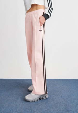 Oseba, ki nosi svetlo roza Adidas trenirke s črnimi črtami, sive superge, z roko v žepu z zadrgo, stoji na modrem preprogi.