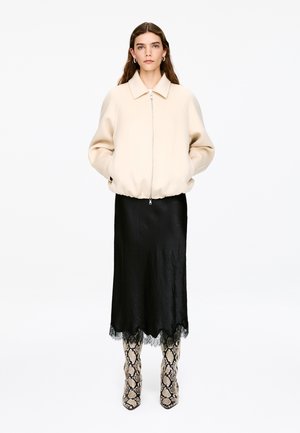 Beige Bomberjacke mit durchgehendem Reißverschluss, schwarzer Satinhauchrock mit Spitzenbesatz und gemusterte kniehohe Stiefel mit Schlangenprint.