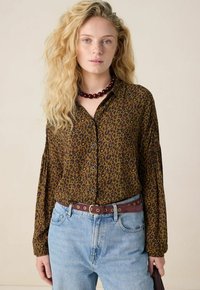 Blouse à imprimé léopard brun avec un design à boutonnage et des manches longues, portée avec un jean bleu clair taille haute et une ceinture bordeaux.