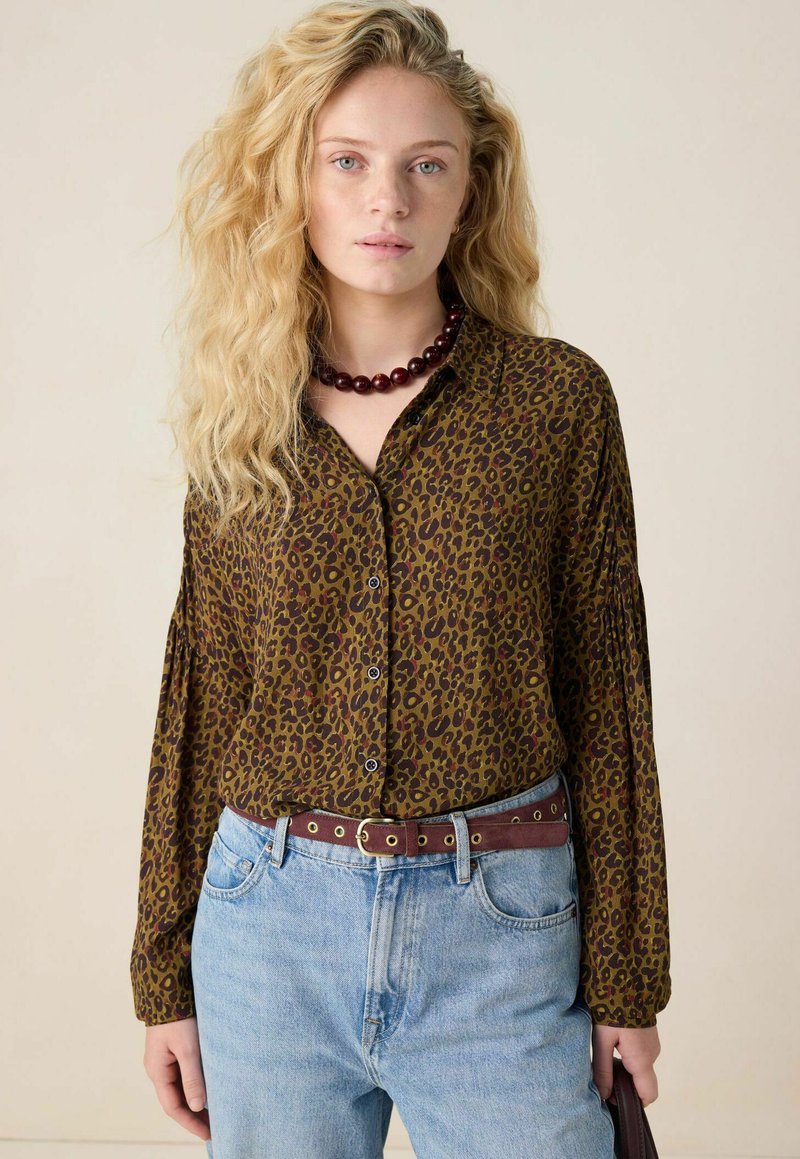 Blouse à imprimé léopard brun avec un design à boutonnage et des manches longues, portée avec un jean bleu clair taille haute et une ceinture bordeaux.
