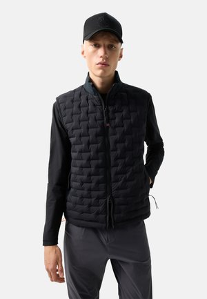 Junger Mann trägt eine schwarze gesteppte Weste, ein schwarzes langärmeliges Shirt, eine schwarze Kappe und dunkelgraue Hose, steht mit einer Hand in der Tasche.