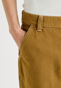 Pantaloni in cotone marrone con design a gamba dritta, caratterizzati da cuciture visibili, tasche laterali e passante per cintura in vita.