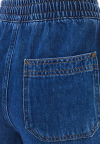 Modré džíny mají elastický pás a zadní kapsu, s kontrastním hnědým prošíváním podél švů a okrajů.