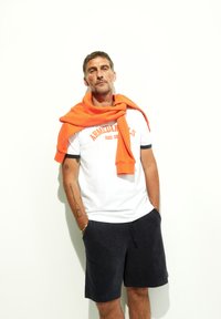 Witte t-shirt met oranje tekst, gecombineerd met marineblauwe shorts. Een oranje sweatshirt is over de schouders gedrapeerd. Soepele stof en casual pasvorm.