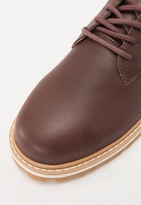 Brown leather shoe with a rounded toe, matching laces, and a textured surface. Features a light-colored stitched sole with traction details.