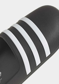 Schwarze Rutschsandale mit drei weißen Streifen auf der Oberseite. Hergestellt aus strukturiertem Material mit leicht geschwungener Form.