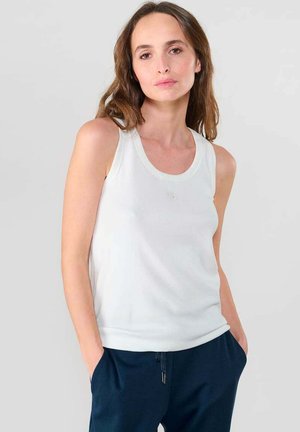 Débardeur blanc à côtes avec un décolleté arrondi et un détail de logo subtil, associé à un pantalon bleu marine, illustrant un design simple et ajusté.