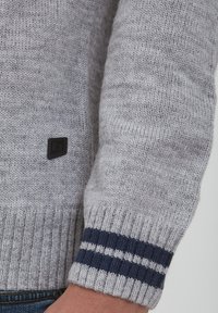 Grå strikket genser med ribbede mansjetter som har marineblå striper, og et lite svart logoklistermerke på siden. Myk tekstur og middels tykkelse.