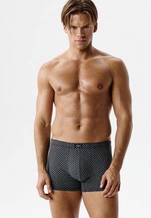 Mężczyzna o umięśnionej sylwetce i krótkich włosach, ubrany w czarne bokserki z wzorem, stojący na jasnym, jednolitym tle.