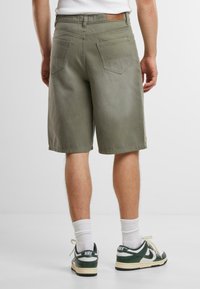 Pantaloni corti da uomo in cotone verde oliva con un design a taglio dritto, tasche posteriori e un'etichetta in pelle sulla vita. Abbinati a sneakers verdi.