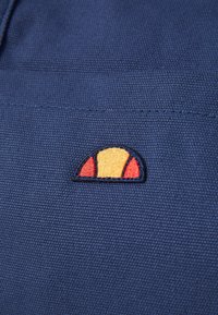 Gros plan sur un tissu bleu texturé avec un petit logo brodé représentant un demi-cercle jaune et rouge cousu.