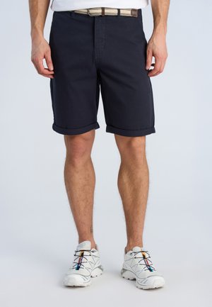 Uomo con pantaloncini blu navy risvoltati, cintura beige intrecciata e scarpe da ginnastica bianche con dettagli multicolore, in piedi su sfondo chiaro.