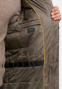 Gepolsterte Jacke mit glänzender olivgrüner Außenseite, gelbem Akzent entlang des Reißverschlusses und einer Innentasche mit Etikett. Weiche Textur.