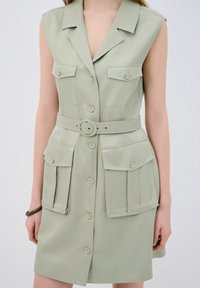 Robe vert clair avec col, design sans manches, deux poches avant et taille ceinturée. Comprend des boutons de fermeture et un tissu texturé.