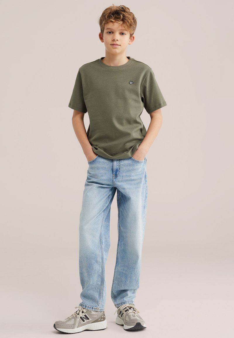 Chlapec s kučeravými vlasmi, oblečený v olivovo zelenej tričku, svetlomodrých džínsach a sivých teniskách, stojaci s rukami v kapsách na neutrálnom pozadí.