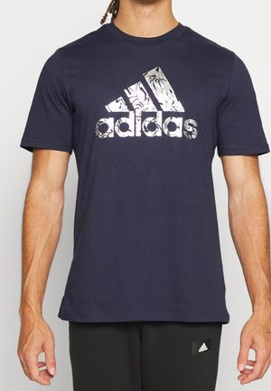 Mann trägt dunkelblaues Adidas-T-Shirt mit silbernem Marmormuster-Logo und schwarze Adidas-Hose, steht vor schlichtem Hintergrund.