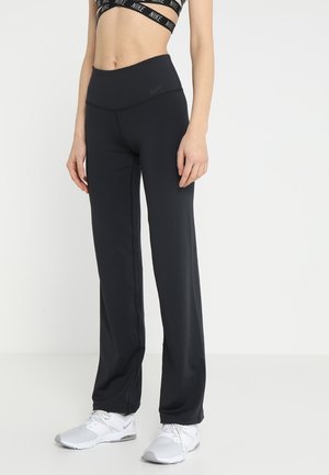 Femme portant un pantalon noir taille haute à jambes larges, un soutien-gorge de sport noir Nike et des baskets blanches Nike, debout devant un fond blanc uni.