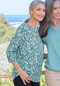 Grüne gemusterte Bluse mit Dreiviertelärmeln, die ein geometrisches Design in Weiß aufweist, kombiniert mit schwarzen Hosen, im Freien.