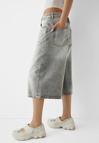 Persona vistiendo una falda de mezclilla descolorida de longitud media, una camiseta corta blanca y zapatillas deportivas blancas con tiras de Velcro, de pie contra un fondo sencillo.