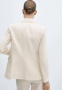 Vrouw met een knot in haar haar draagt een lichtbeige blazer en bijpassende broek, staande tegen een effen witte achtergrond, met haar gezicht van ons af.