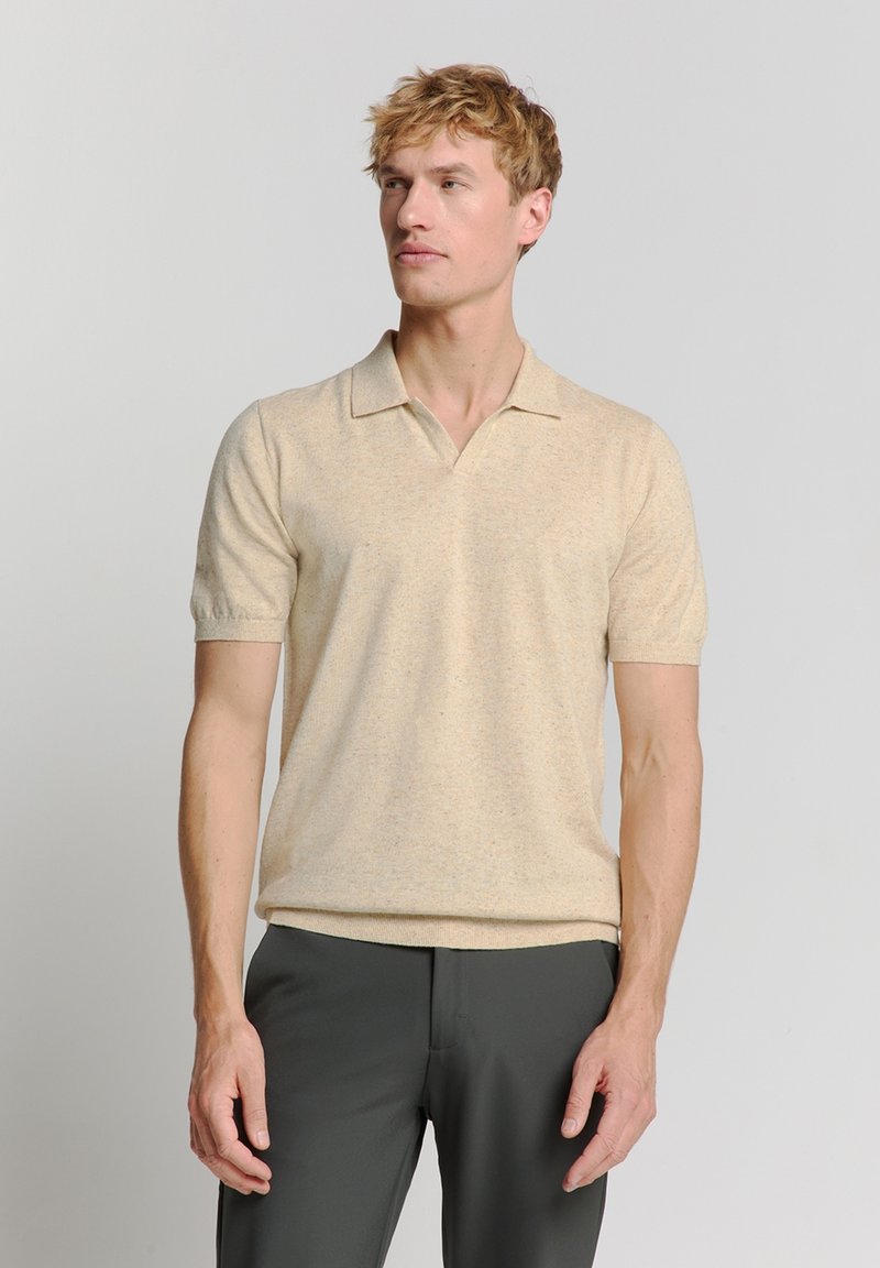Beige polo shirt met korte mouwen, gemaakt van een zachte, gestructureerde stof met een klassieke kraag en een subtiel gespikkeld patroon. Gecombineerd met donkere broeken.
