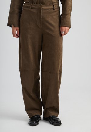 Person som bär bruna byxor med vida ben och svarta blanka loafers, med händerna vilande vid sidorna och silverringar på fingrarna.