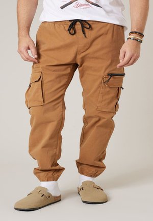 Homme portant un pantalon cargo marron avec des chevilles élastiques, des chaussettes blanches, des sandales beige à enfiler et un t-shirt blanc avec un motif, ainsi que des bracelets au poignet droit.