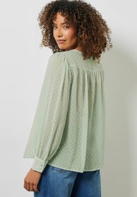 Hellgrüne Bluse mit transparenter Textur, Gepunktetes Muster, Puffärmel und Knopfmanschetten, kombiniert mit blauen Jeans.