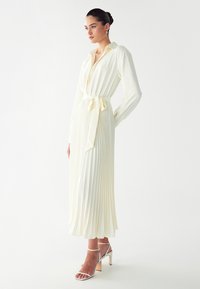 Langes, plissiertes Kleid aus weichem Creme-Stoff mit einem kragenähnlichen Ausschnitt. Verfügt über einen seitlichen Bindegürtel und lange Ärmel, kombiniert mit Riemchen-Heels.