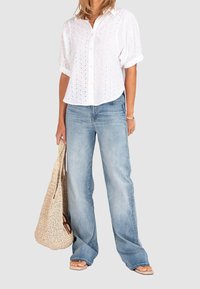 Camicia bianca con dettagli in pizzo, abbinata a jeans azzurri svasati. Borsa tote intrecciata beige, sandali flat, stile casual.