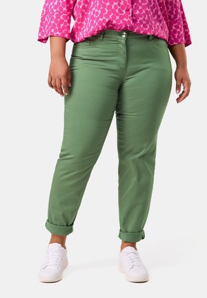 Grüne Hose mit umgeschlagenen Bündchen, schlankem Design und glatter Textur, kombiniert mit einem gemusterten pinken Oberteil und weißen Turnschuhen.