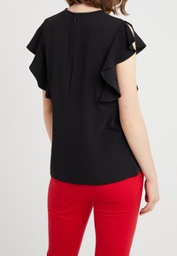 Kvinna som bär en svart ärmlös topp med volangärmar och röda byxor, sedd bakifrån mot en plain vit bakgrund.