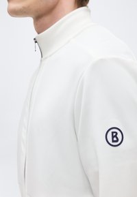 Άνδρας που φορά λευκό μπουφάν με φερμουάρ και όρθιο γιακά, με μαύρο κυκλικό λογότυπο που περιέχει ένα "B" στο αριστερό πάνω μανίκι.