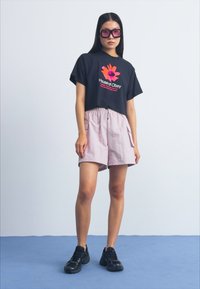 Tricou scurt negru cu logo-ul "House of Obey", asortat cu pantaloni scurți roz deschis. Modelul poartă adidași negri și ochelari de soare roz supradimensionați.