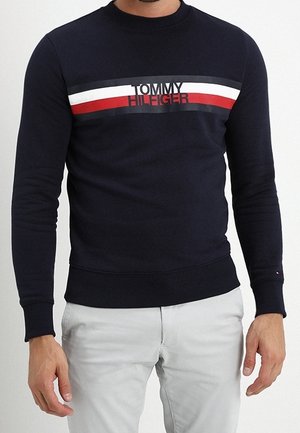 Mężczyzna w granatowym swetrze Tommy Hilfiger z białymi, czerwonymi i czarnymi poziomymi paskami na piersi, w połączeniu z jasnoszarymi spodniami.