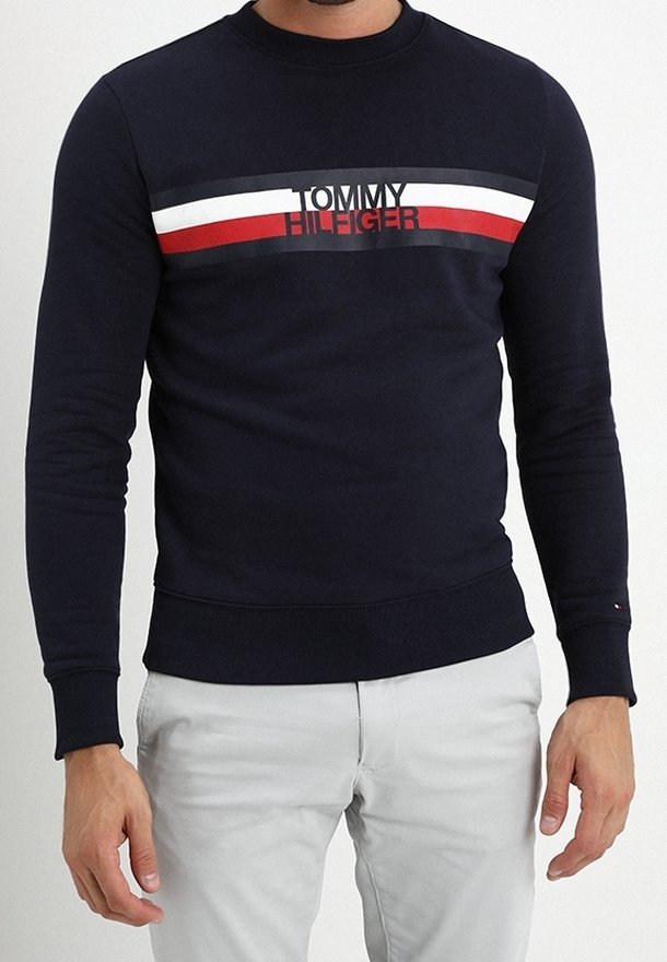 Homme portant un pull Tommy Hilfiger bleu marine avec des rayures horizontales blanches, rouges et noires sur la poitrine, associé à un pantalon gris clair.