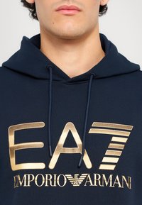 Mørkeblå hettegenser med gull "EA7 EMPORIO ARMANI" logo. Har snøring i hetta og ribbestruktur. Enkel, sporty design.
