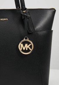 Sac à main en cuir noir texturé avec une fermeture éclair dorée, une poche latérale, et un pendentif circulaire doré avec le logo MK accroché à la poignée.