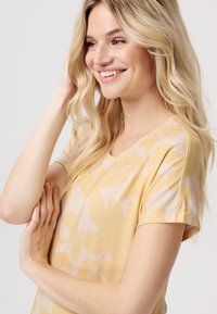 Lichtgele top met korte mouwen en een bloemenpatroon, met een ronde hals en een zachte textuur. De hand van het model raakt haar haar aan in een casual pose.