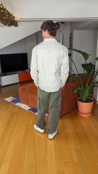 Camisa de botones a rayas, pantalones cargo color oliva y zapatillas blancas. Sofá marrón texturizado y planta verde en maceta en una habitación bien iluminada. Suelo de madera.