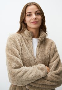 Beige fleecetrui met hoge kraag, ritssluiting, zachte textuur en geen zichtbare patronen. Draagt over een wit overhemd.