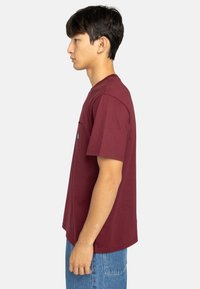 Camiseta burdeos de algodón con manga corta, cuello redondo y un bolsillo en el pecho que presenta una pequeña etiqueta; combinada con unos jeans de color azul claro.