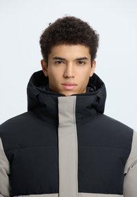 Polstret jakke med høj krave, der har et farveblokdesign i sort og beige. Fremstillet af glat stof med en mat finish.
