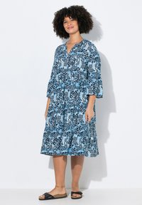 Blaues Kleid mit Blumenmuster, 3/4-Ärmeln, V-Ausschnitt und gestuften Design. Weicher Stoff und lässige Passform, getragen mit schwarzen Slipper-Sandalen.
