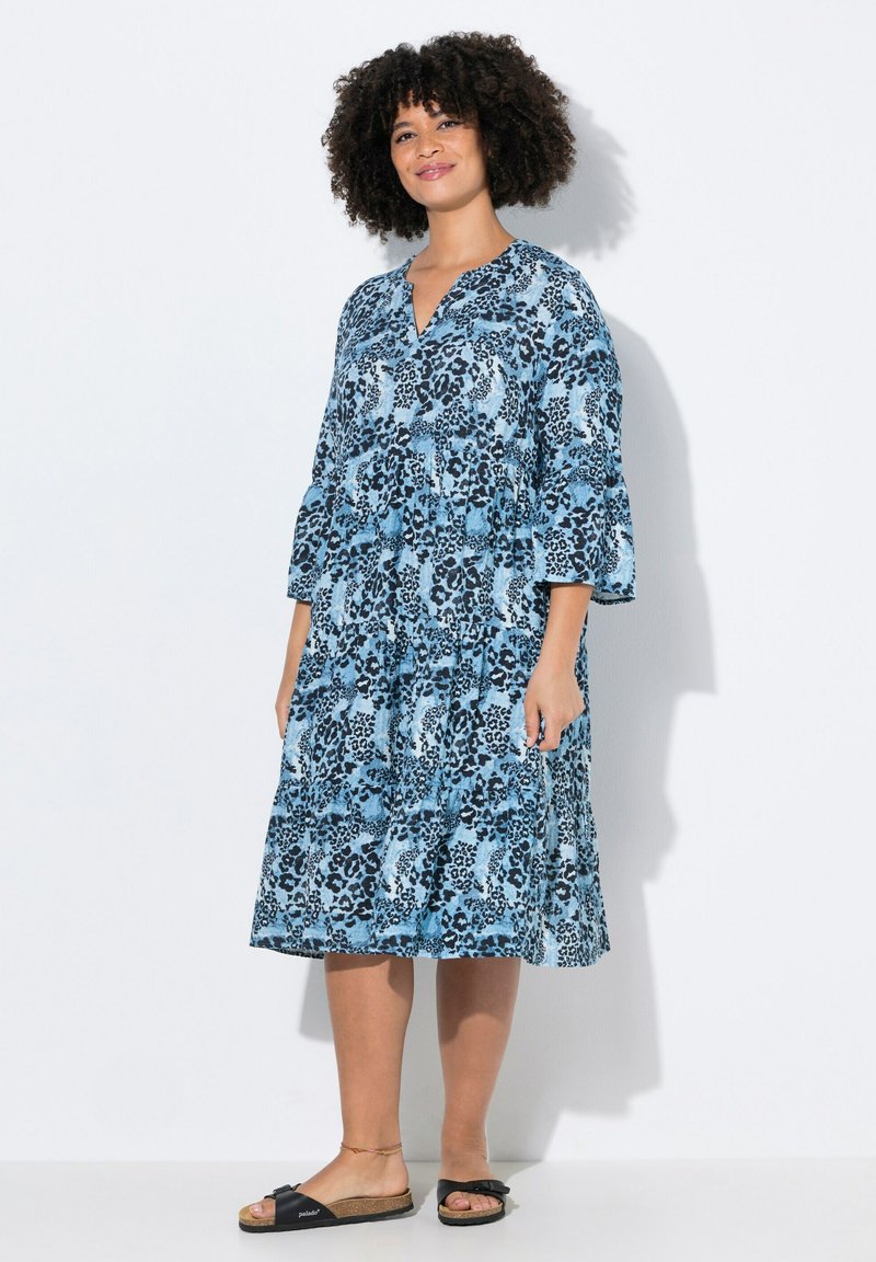 Blaues Kleid mit Blumenmuster, 3/4-Ärmeln, V-Ausschnitt und gestuften Design. Weicher Stoff und lässige Passform, getragen mit schwarzen Slipper-Sandalen.