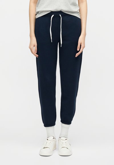 Άτομο που φοράει σκούρο μπλε παντελόνι jogger με λευκό κορδόνι, σε συνδυασμό με λευκές κάλτσες και λευκά αθλητικά παπούτσια, στέκεται μπροστά σε απλό φόντο.