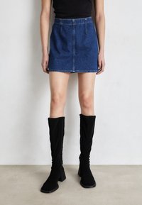 Modell iförd en blå denim minikjol, svart ärmlös topp och svarta knähöga klackstövlar som står på ett enhetligt golv.