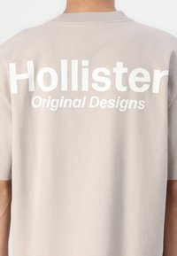 Hellgraues Baumwoll-T-Shirt mit großen weißen Buchstaben auf dem Rücken: "Hollister" über "Original Designs." Kurzarmdesign und Rundhalsausschnitt.