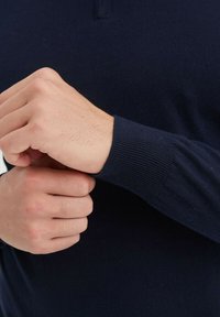 Pull en tricot bleu marine avec un poignet côtelé, présentant un col montant et une texture fine, exhibé avec des mains ajustant la manche.