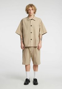 Beige kortermet skjorte med brystlomme og svarte knapper, kombinert med matchende beige shorts, brukt med hvite sokker og svarte loafers.